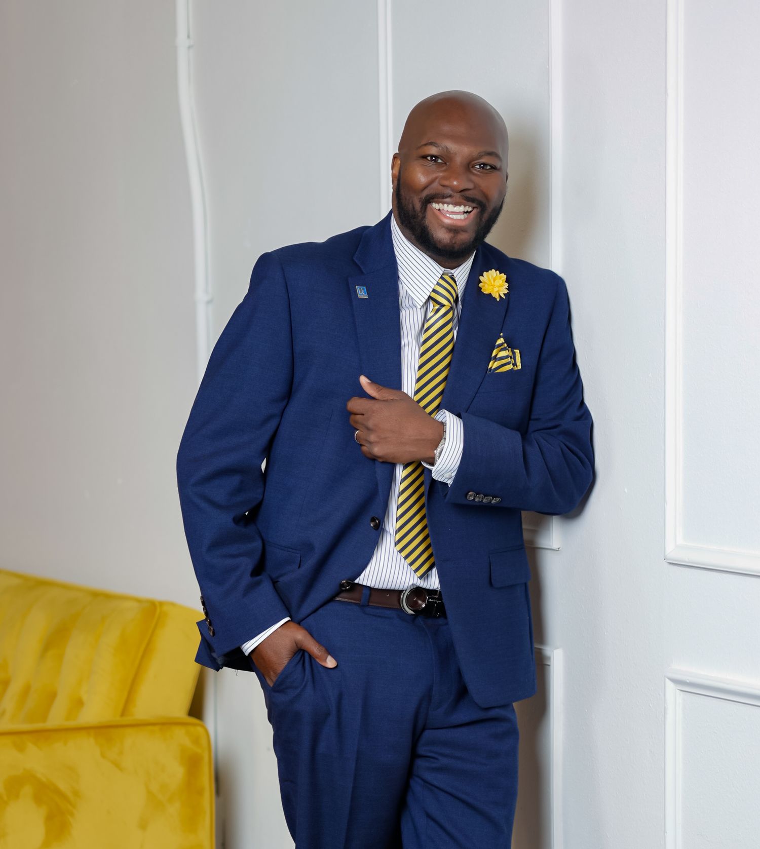 A smiling man in a navy blue suit with yellow accessories poses confidently indoors.