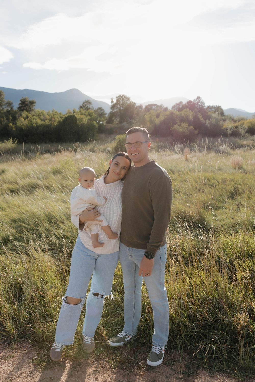 Family walking together through a sunlit meadow with tall grass and mountains in the background at golden hour.