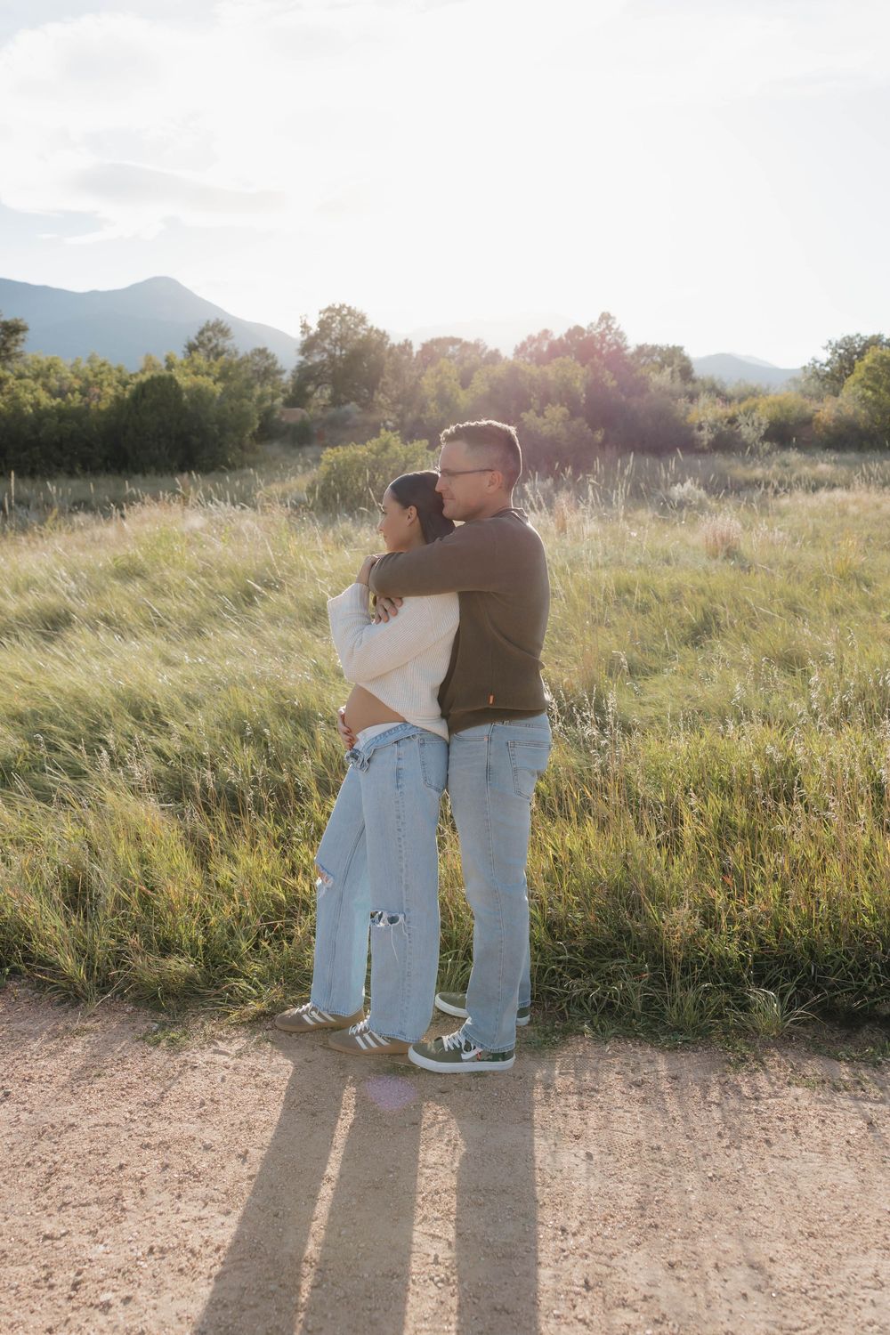 A couple embraces while standing on a dirt path surrounded by tall grass and mountains at golden hour.