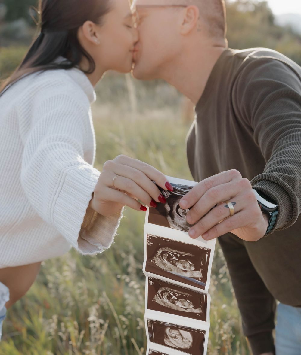 A couple sharing an intimate moment while holding ultrasound photos in a sunny outdoor field.