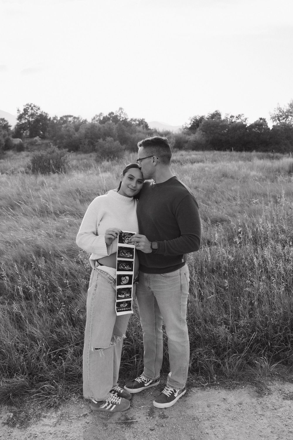 A couple shares a tender moment in a field while holding an ultrasound photo in black and white.