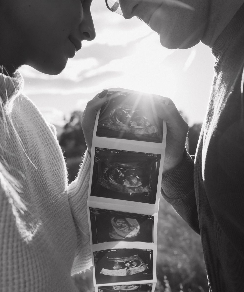Black and white photo of couple looking at ultrasound images together in sunlight, creating a tender pregnancy announcement moment.