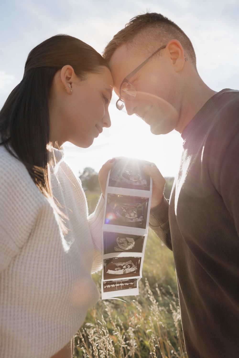 A couple shares a tender moment looking at ultrasound photos together in a sunlit field at golden hour.