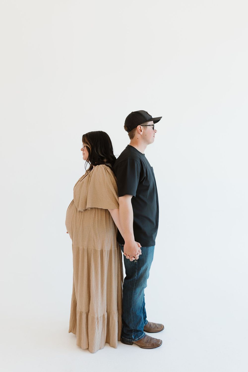 A couple stands back-to-back holding hands against a white background, the woman in tan dress and man in dark clothing.