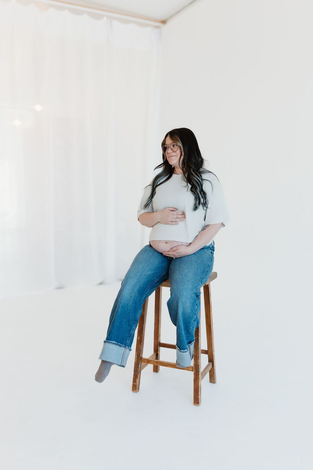 Pregnant woman in denim and grey top sits on wooden stool in white minimalist setting.