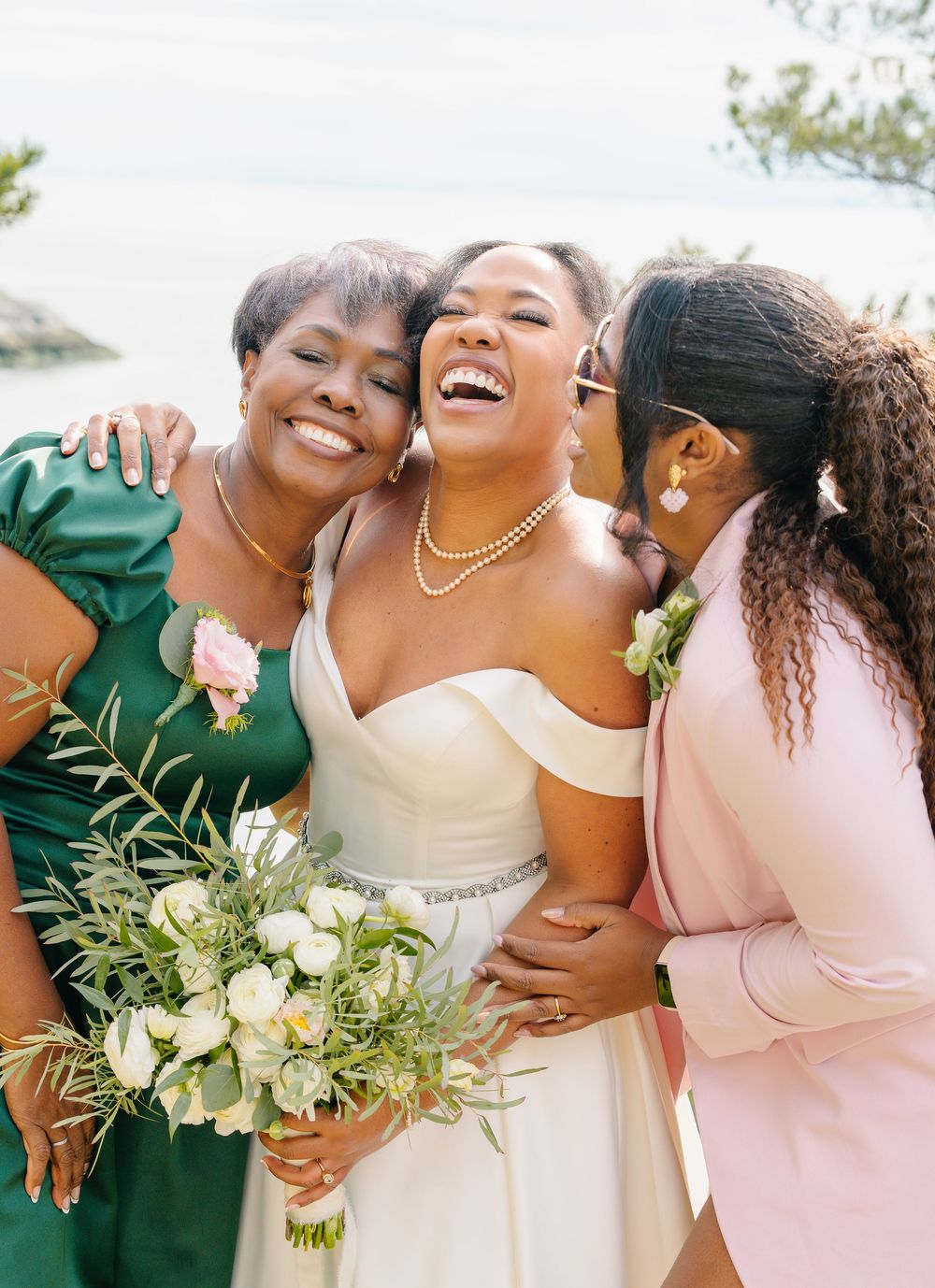 Three people share a joyful embrace at a wedding while wearing elegant dresses and holding a white floral bouquet.