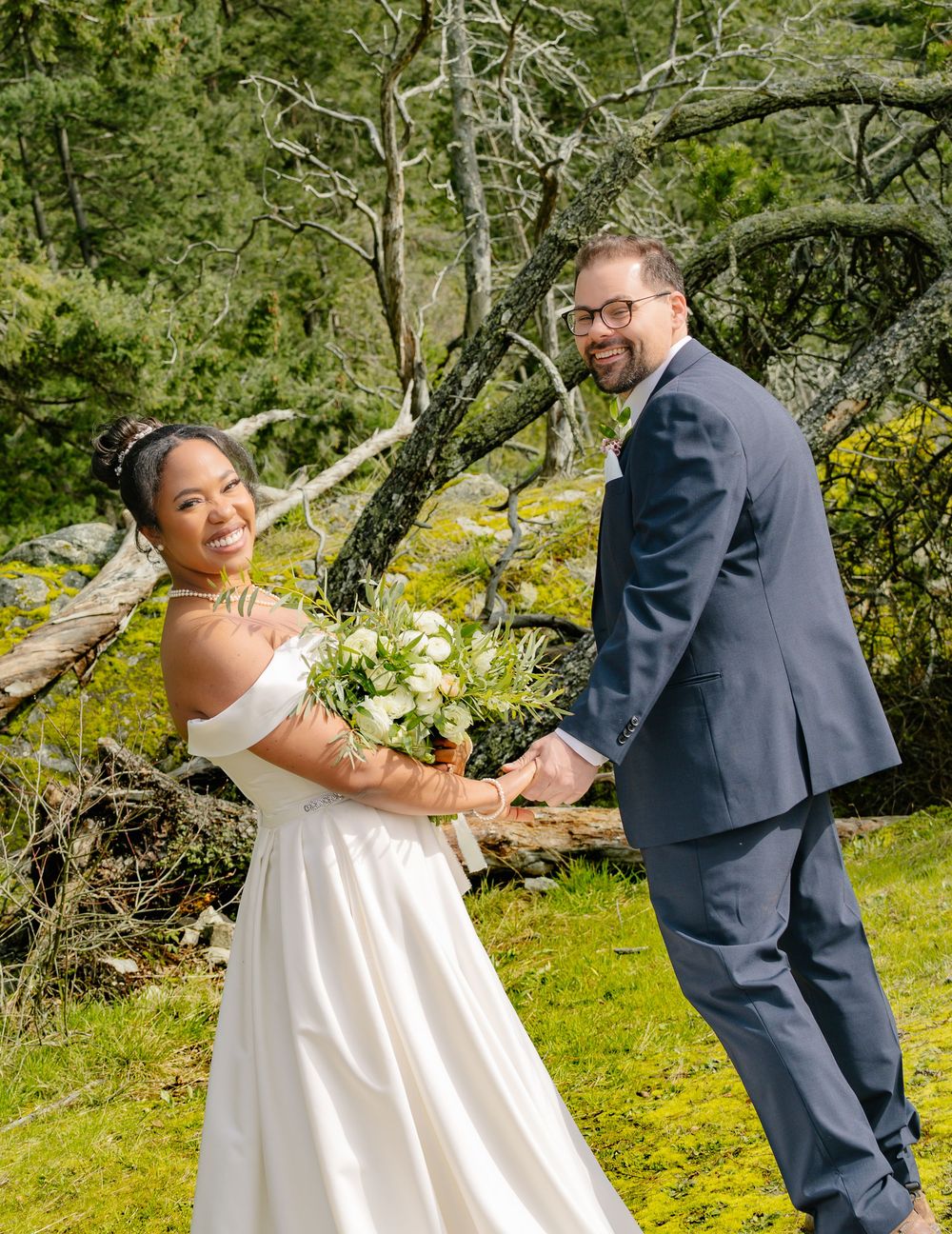 Two people in wedding attire pose together outdoors in a garden setting with blooming flowers and greenery.