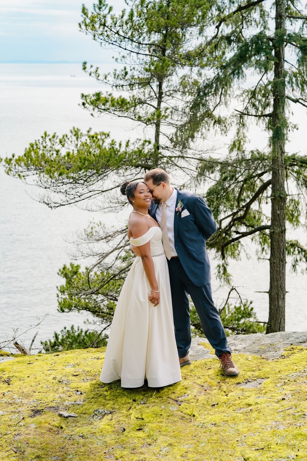 Couple shares romantic moment on mossy cliff overlooking misty coastline with evergreen trees framing the scene.