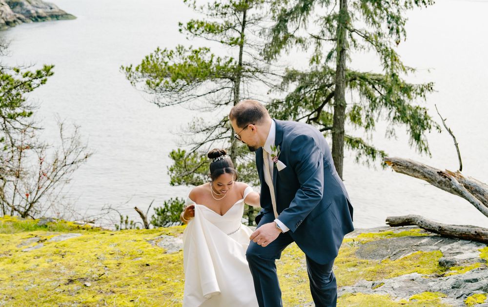 A joyful wedding couple shares a tender moment on a mossy waterfront cliff overlooking the ocean on their special day.
