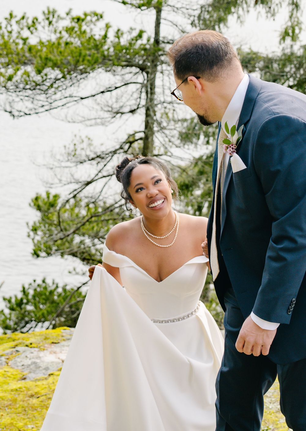 A joyful wedding couple shares a candid moment outdoors against evergreen trees and moss-covered rocks.