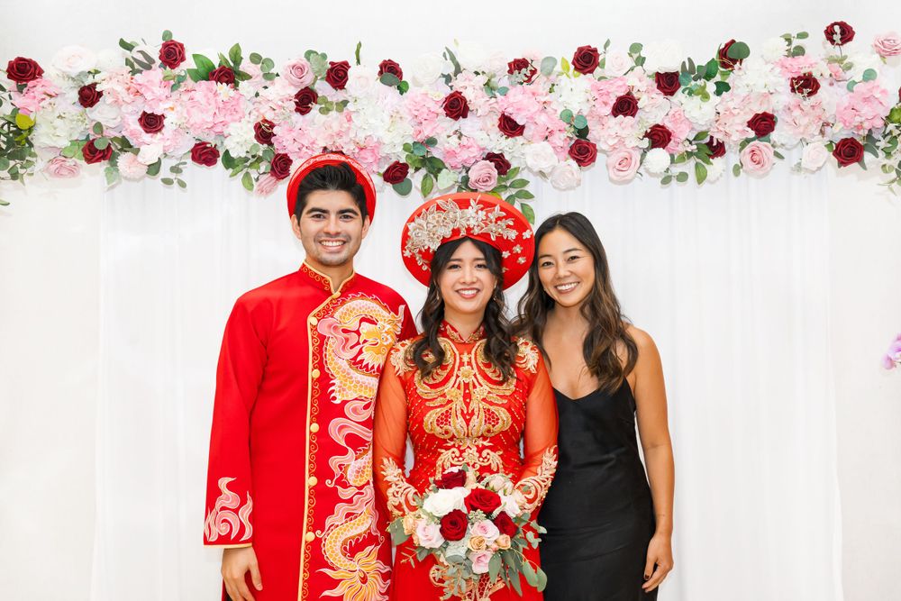 Wedding party poses in front of floral arch with couple in traditional red Vietnamese wedding attire.