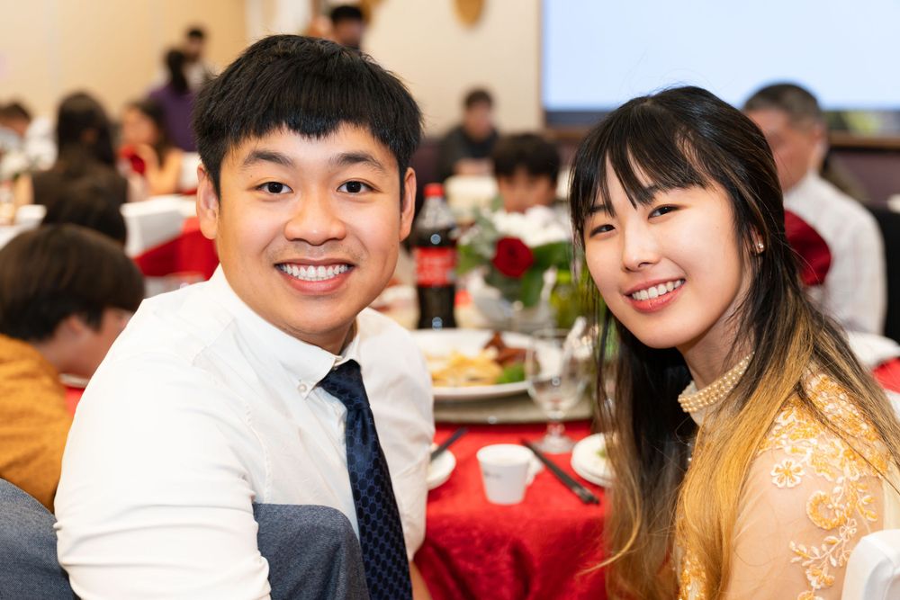 Wedding reception guests in formal attire smiling at dinner table with red decor.