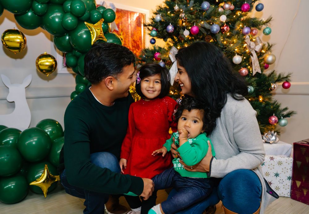 Family celebrates Christmas together sitting by decorated tree with green balloon arch and festive ornaments.