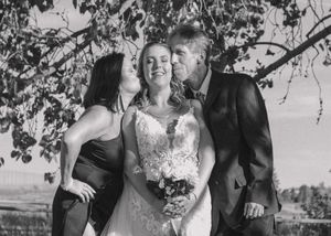 Black and white photo of bride receiving kisses on each cheek from her mother and father, capturing a loving moment.
