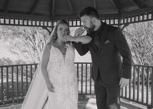 Black and white photo of bride and groom posing in a gazebo, arms intertwined, drinking champagne.