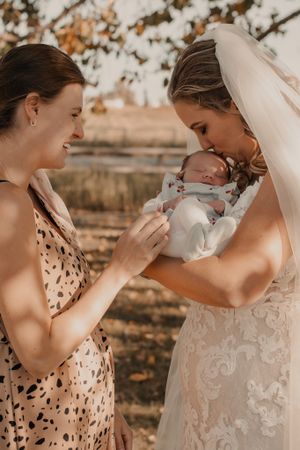 Candid moment of bride gently kissing her sister's newborn on the forehead outside, capturing love and tenderness.