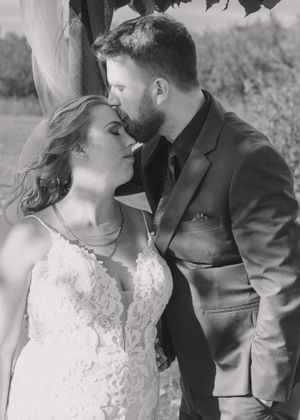 Groom kisses bride's forehead in a tender moment, captured in classic black and white.