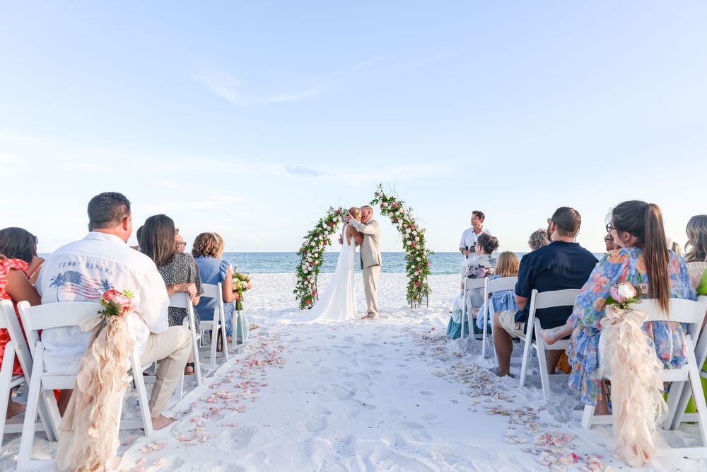 Beachside wedding ceremony setup with white chairs and floral arch against turquoise waters.