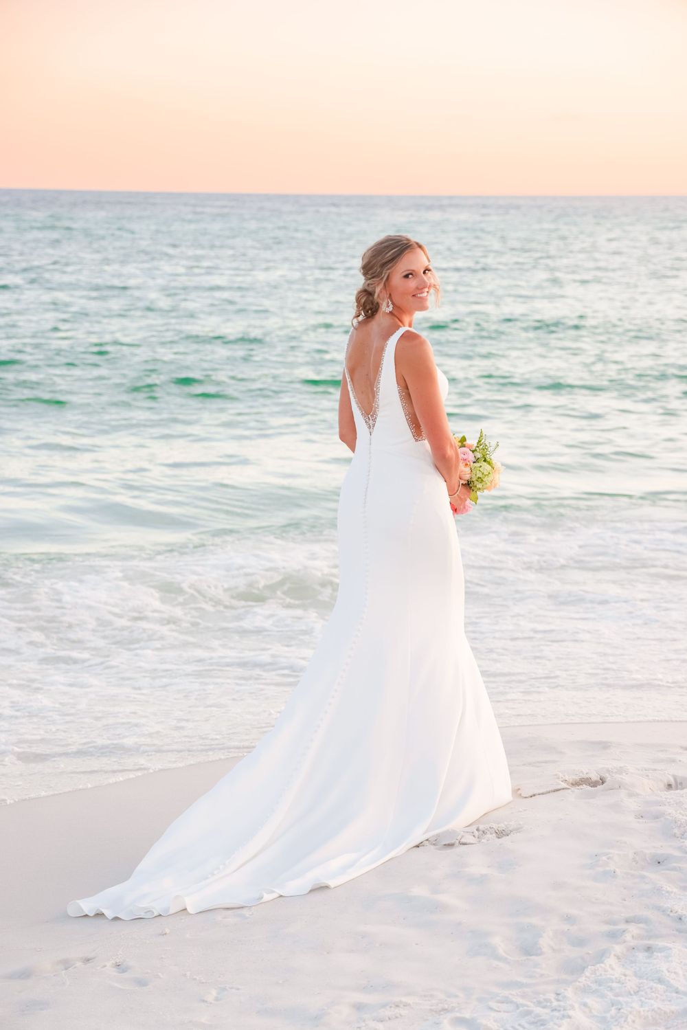 Elegant bride in flowing white dress poses on beach during sunset with bouquet.