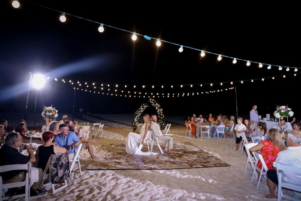 String lights illuminate a nighttime beach wedding reception with guests seated at tables.