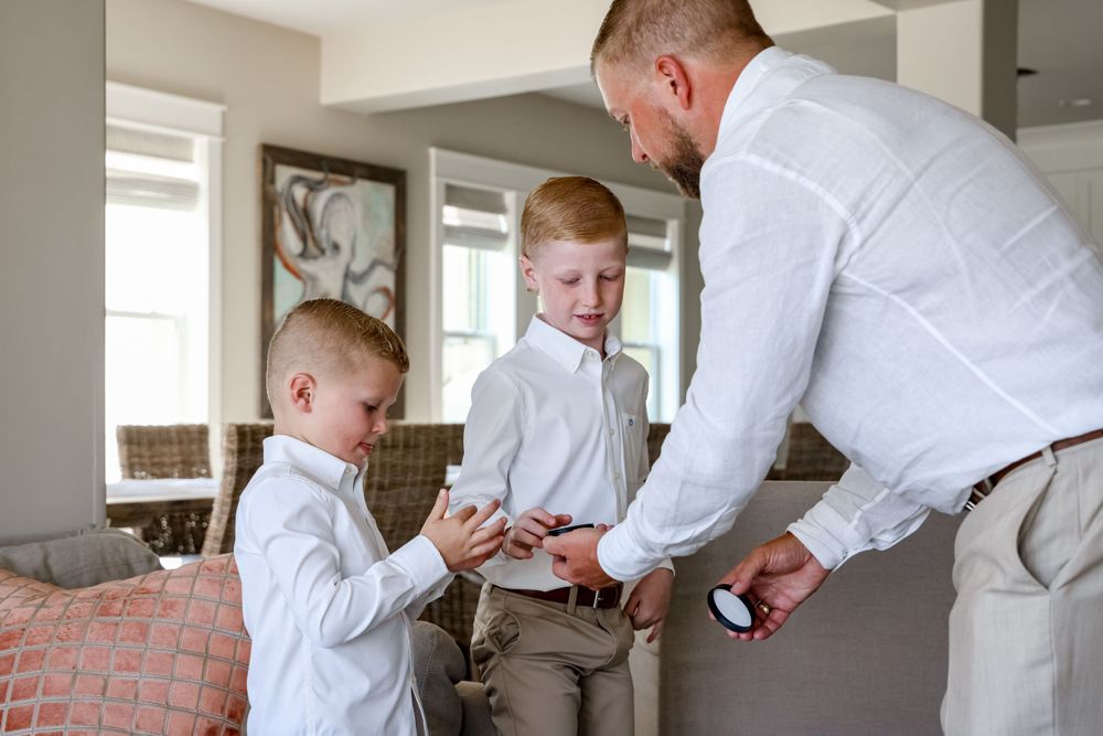 Two young boys in light colored clothing interact with an adult wearing a white shirt indoors.