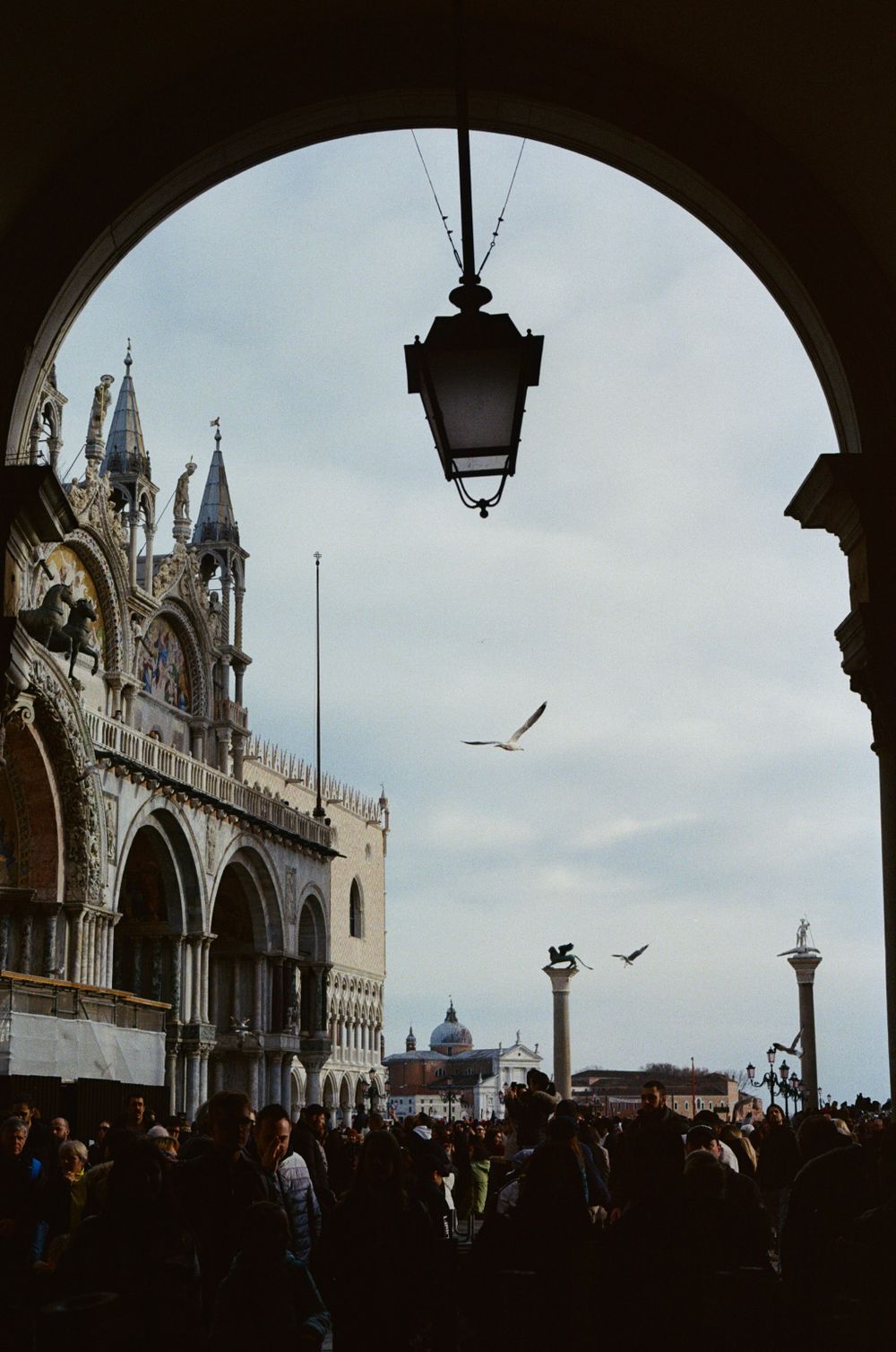 Iconic view of St Mark's Basilica in Venice with silhouetted lantern and flying birds against cloudy sky.