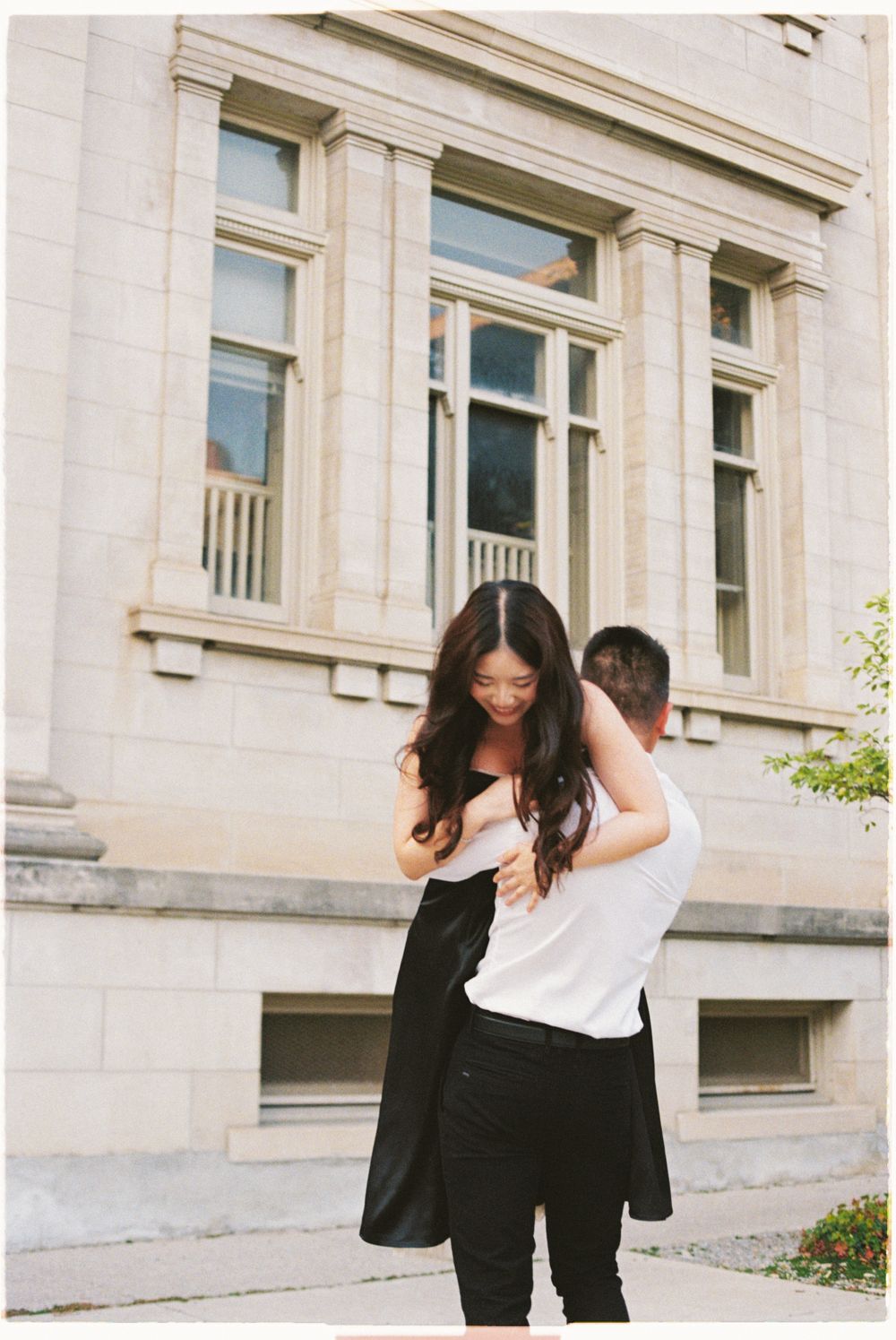 A couple embracing and laughing together in front of a classical stone building.