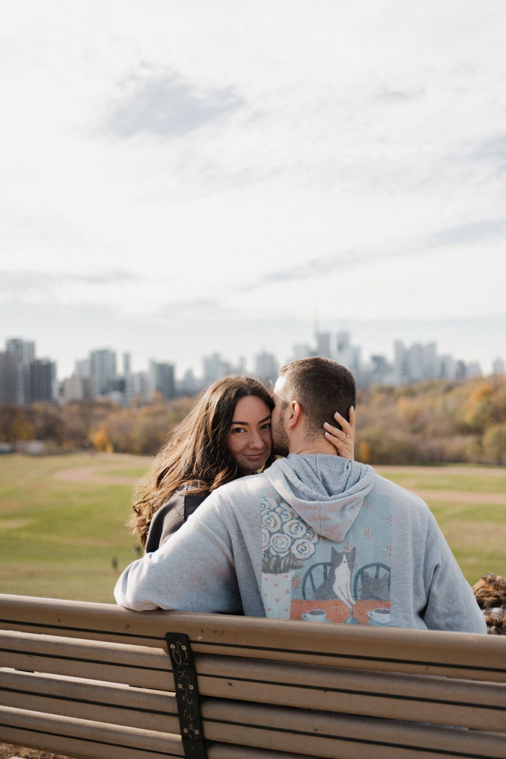Couple sharing intimate moment while sitting on park bench with city skyline in background.