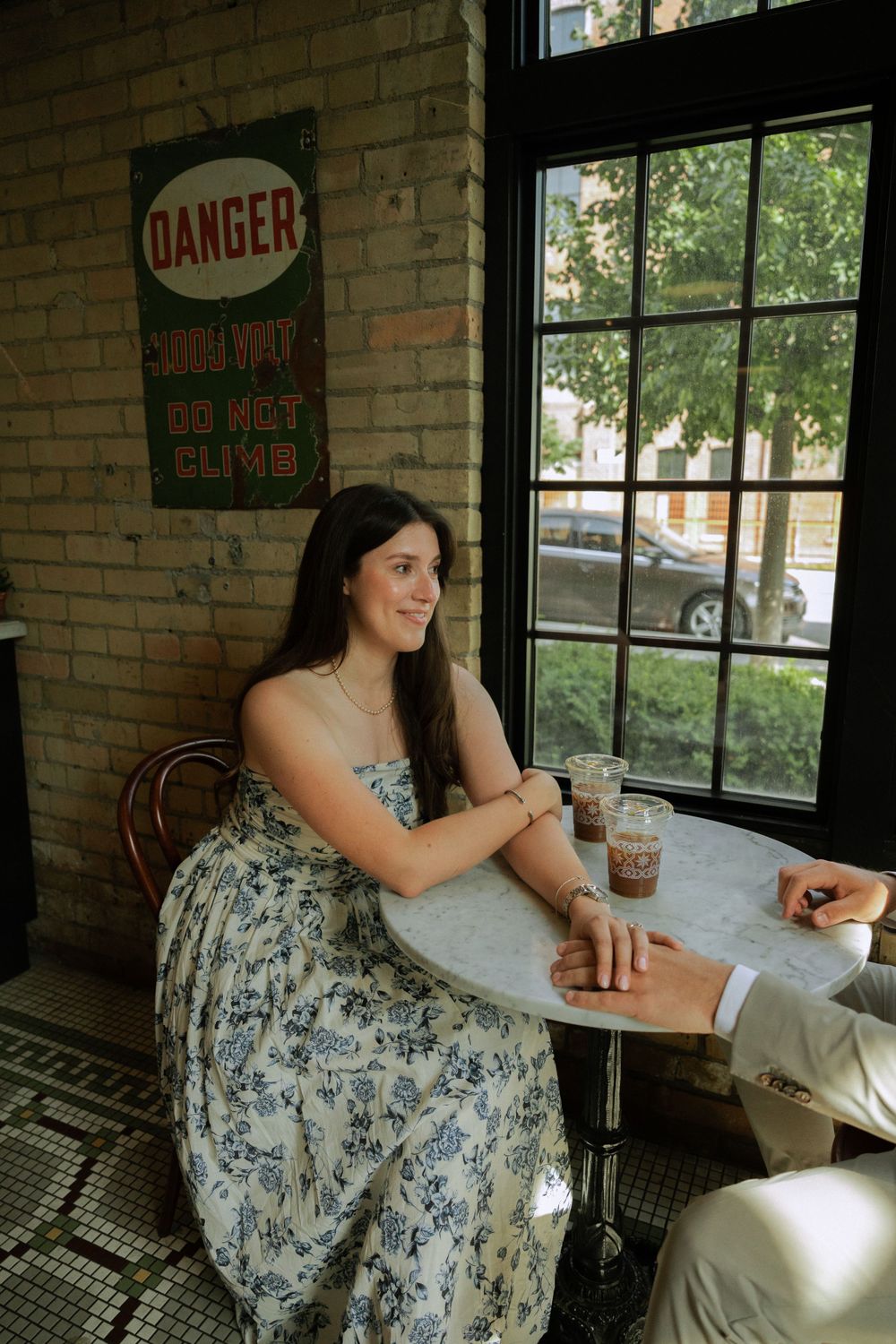 A woman in a floral dress sits at a cafe table near a brick wall with a danger sign.
