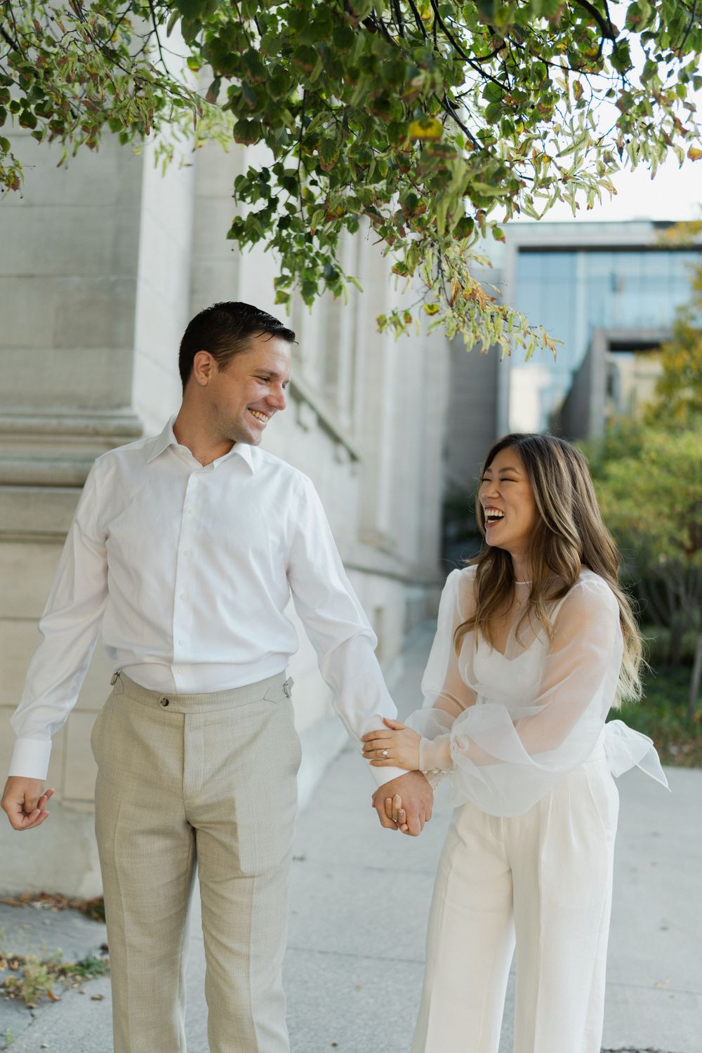 A couple laughing together outdoors in all-white outfits near a classical building.