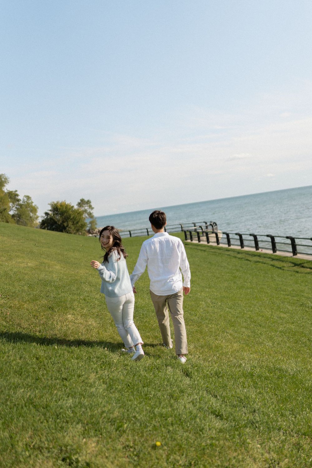 A couple walks together on green grass along a waterfront path on a sunny day.