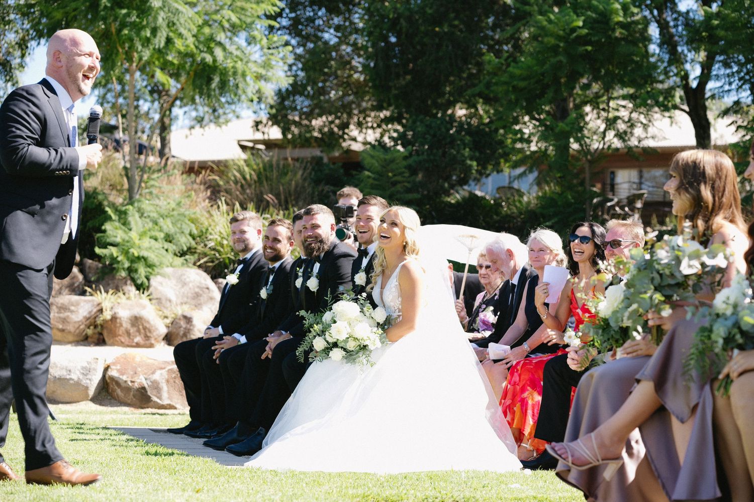 An outdoor wedding ceremony with guests seated on chairs among garden landscaping.