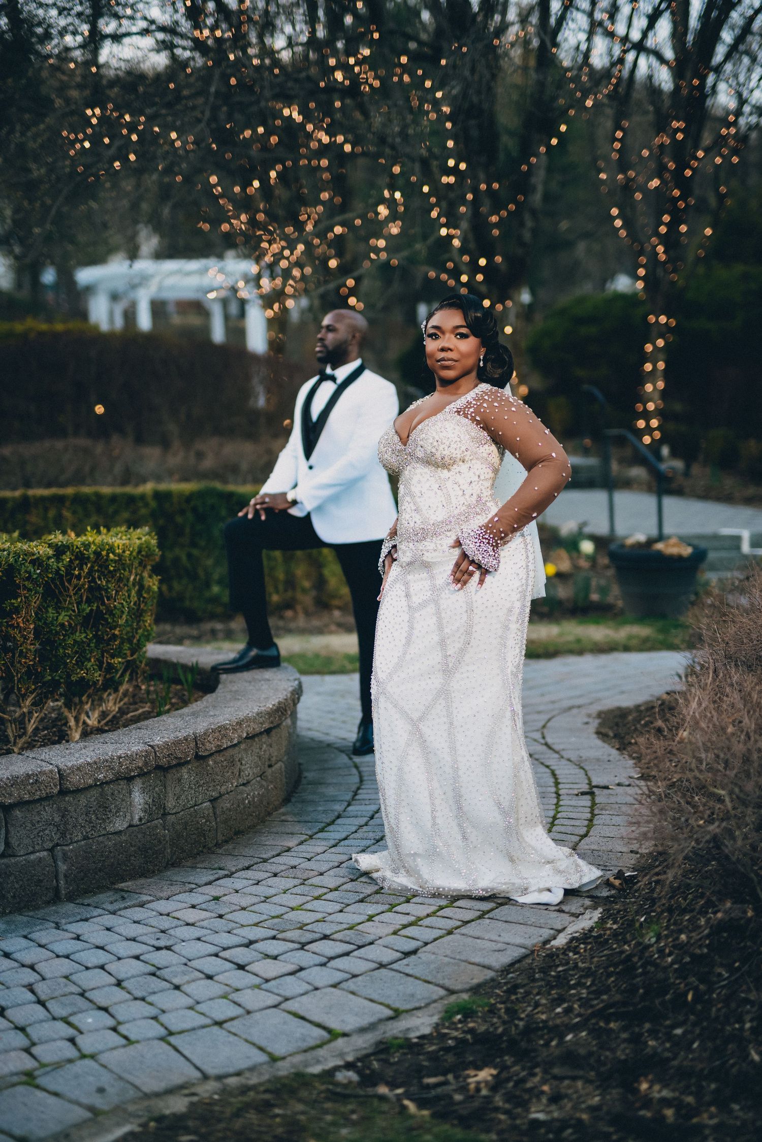 A couple poses elegantly on a stone pathway with glowing string lights in the background during a wedding celebration.
