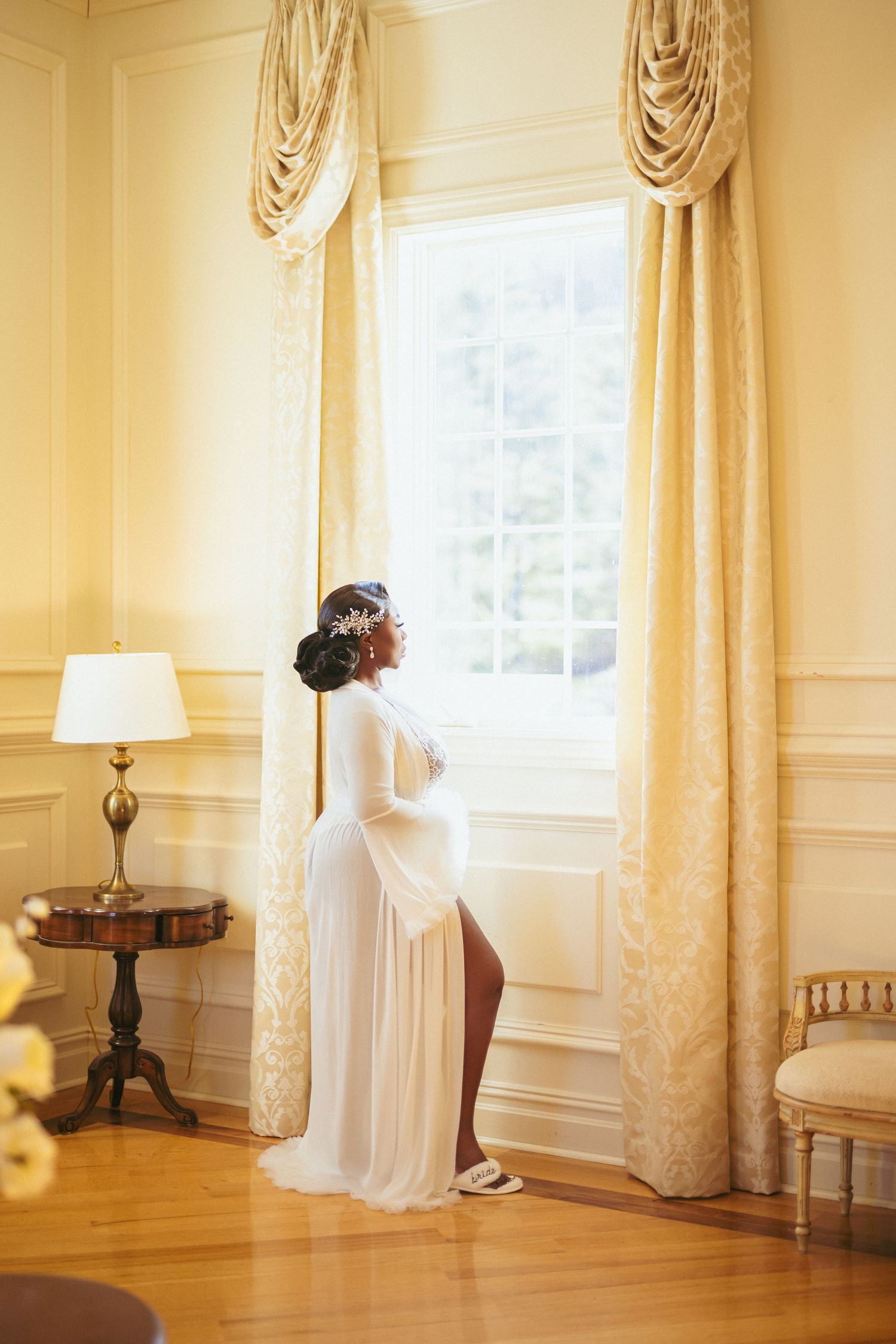 Elegant white flowing maternity gown silhouetted against a bright window with cream curtains in a luxurious room.