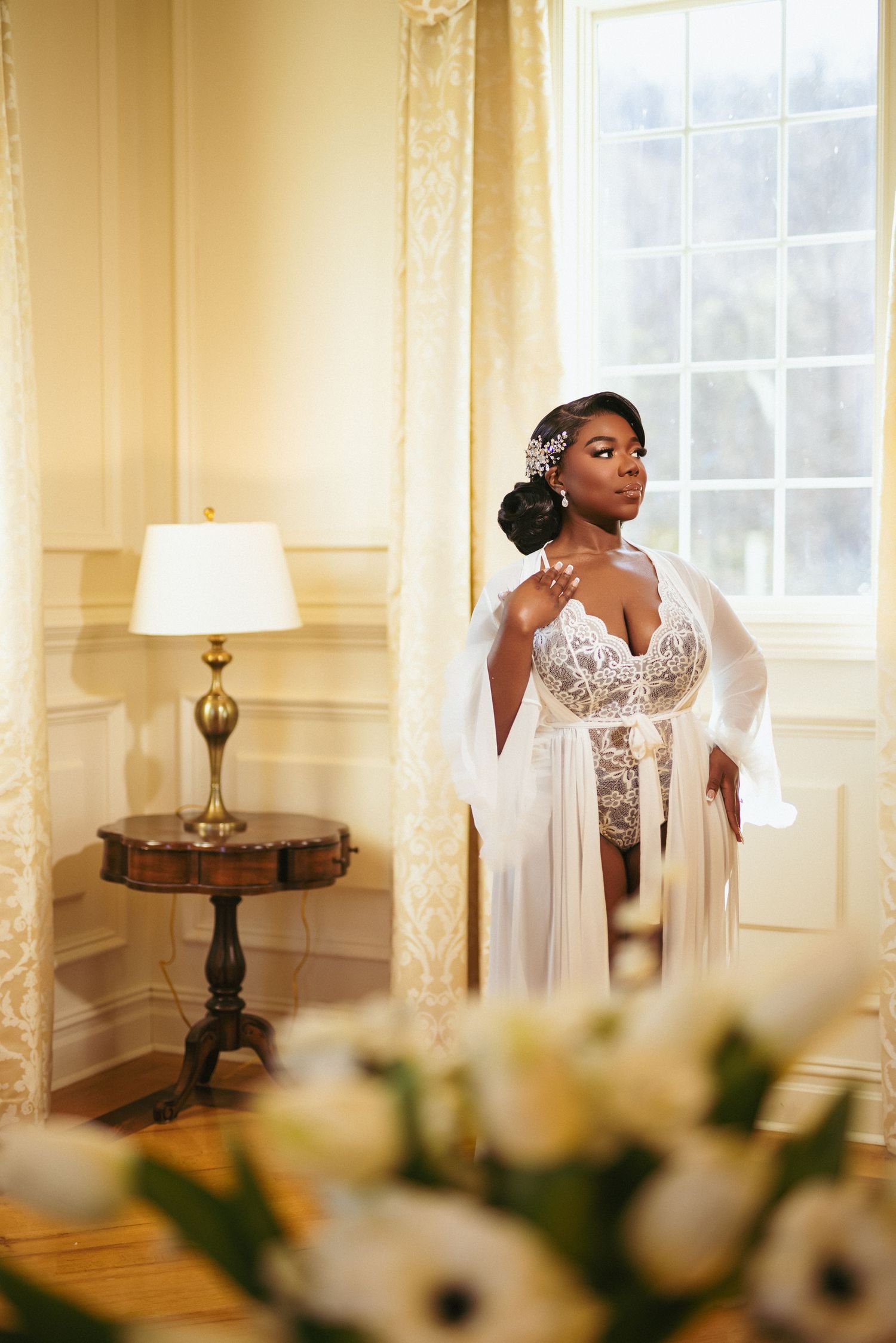 Elegant bride in white lace gown and flowing cape poses by window in luxurious room with antique lamp and floral decor.