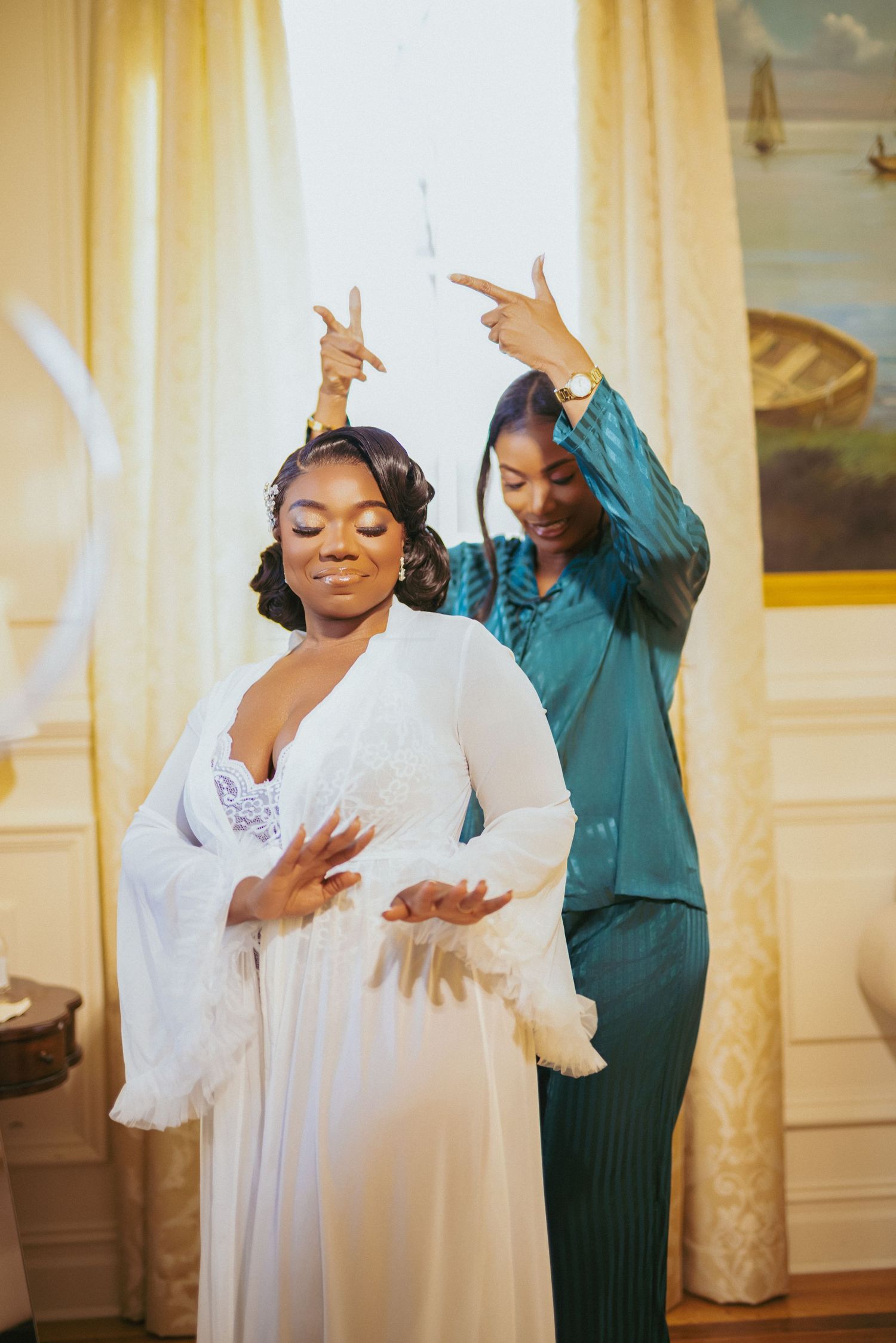Two women share a joyful moment while dancing together, one in a flowing white dress and another in teal attire.