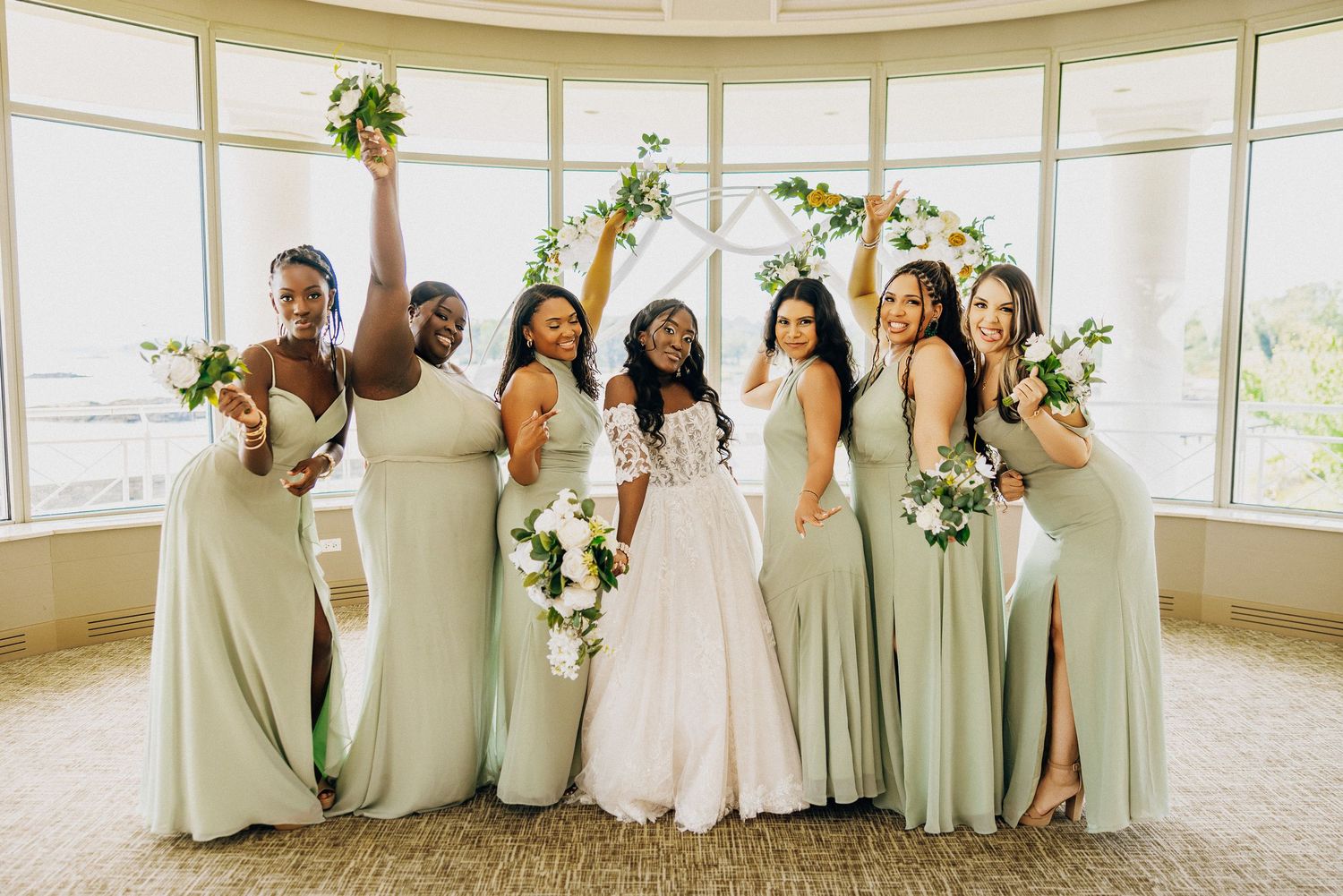 Bridesmaids in sage green dresses and bride in white pose together in front of curved window wall.