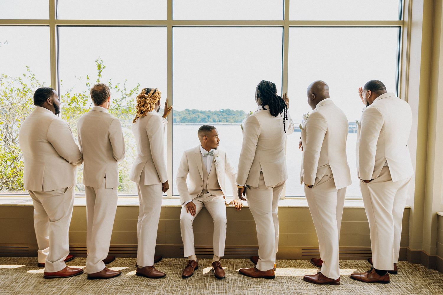Wedding party members in white suits stand in line looking out large windows in an elegant venue.