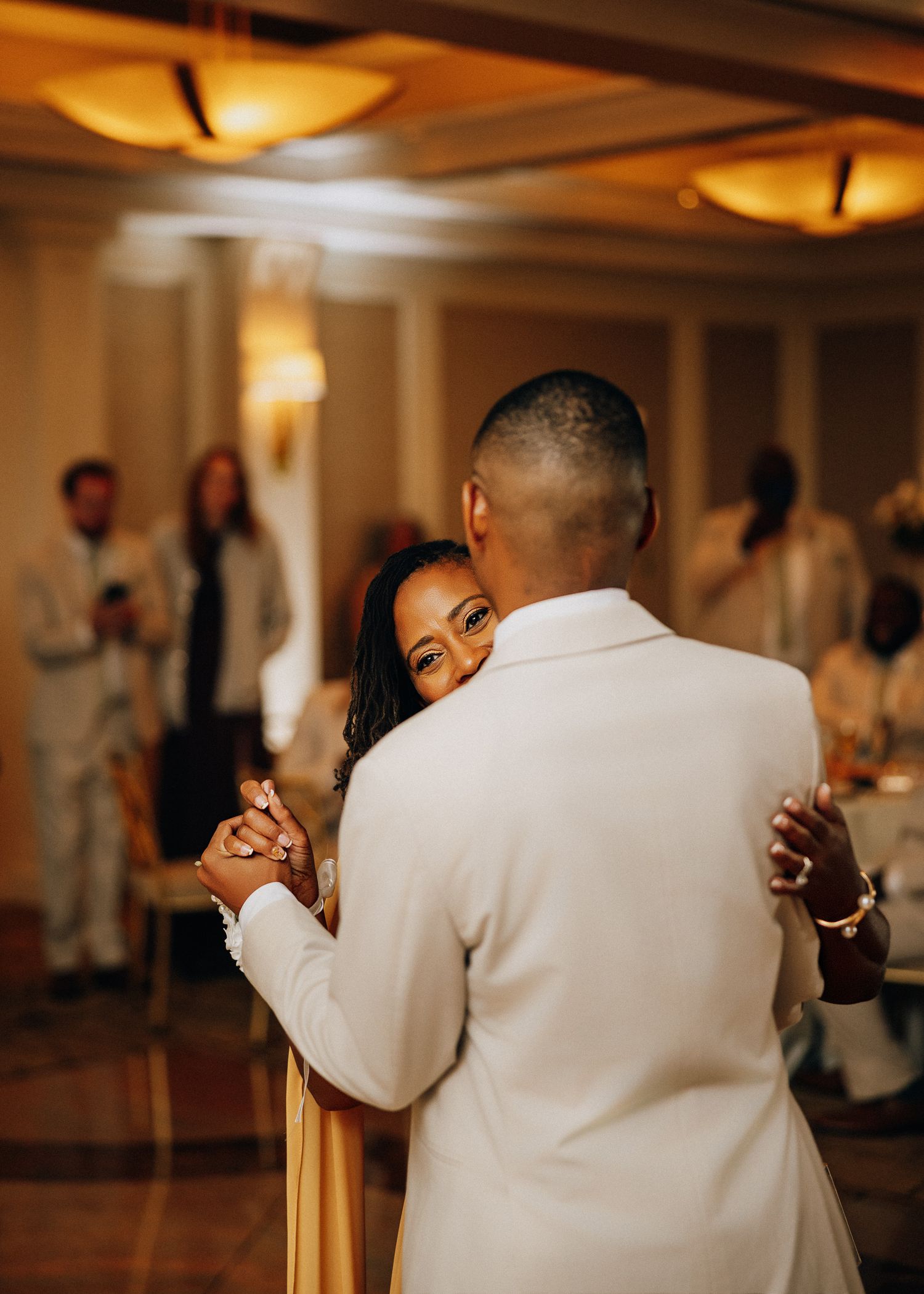 A couple shares an intimate slow dance at their wedding reception, with warm lighting and guests in the background.
