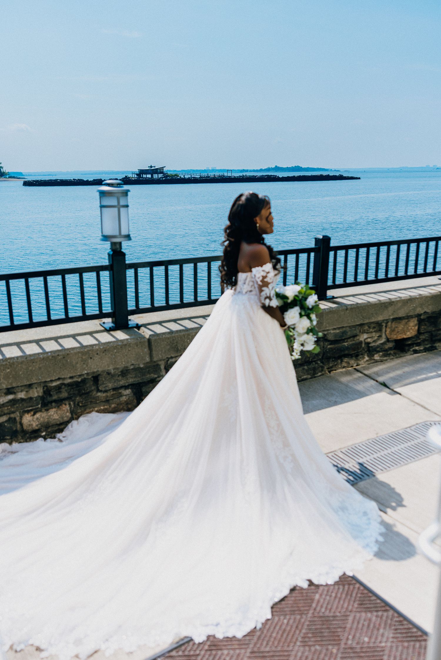 A flowing white wedding dress with a long train photographed against an oceanfront railing on a bright sunny day.