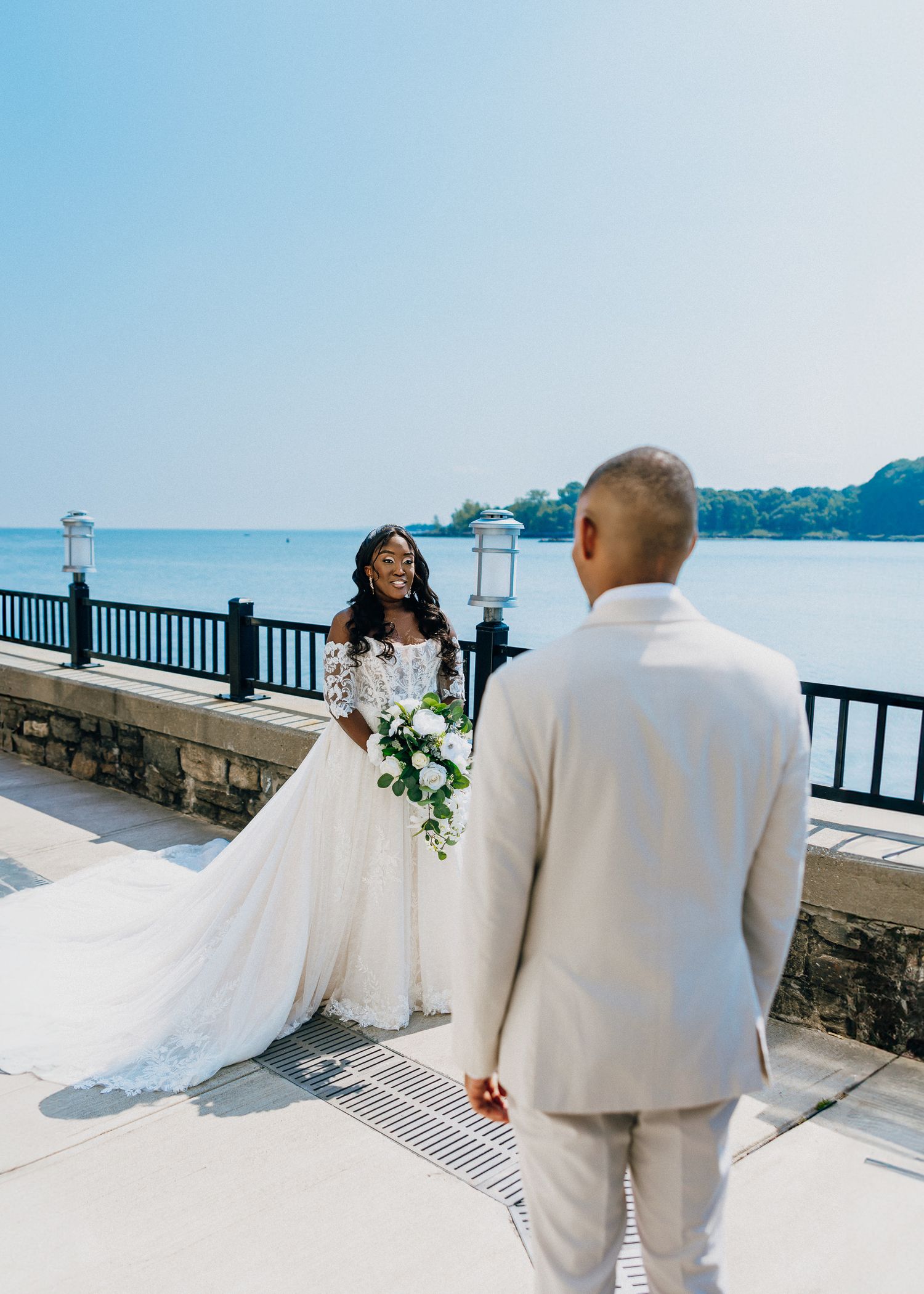 A couple in white attire walks along a scenic waterfront railing on their wedding day.