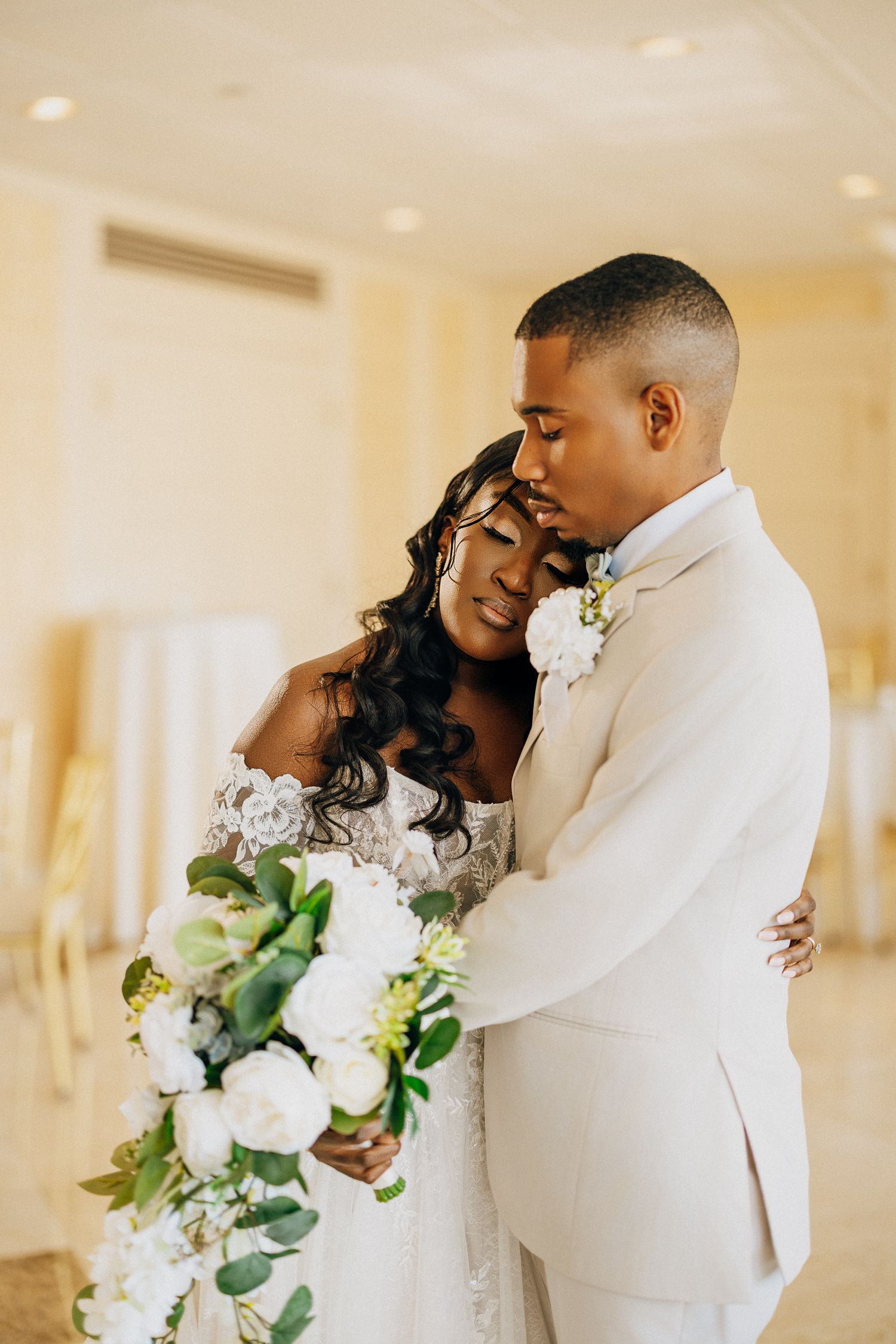 A romantic wedding couple embraces in an elegant white venue while holding a beautiful floral bouquet.