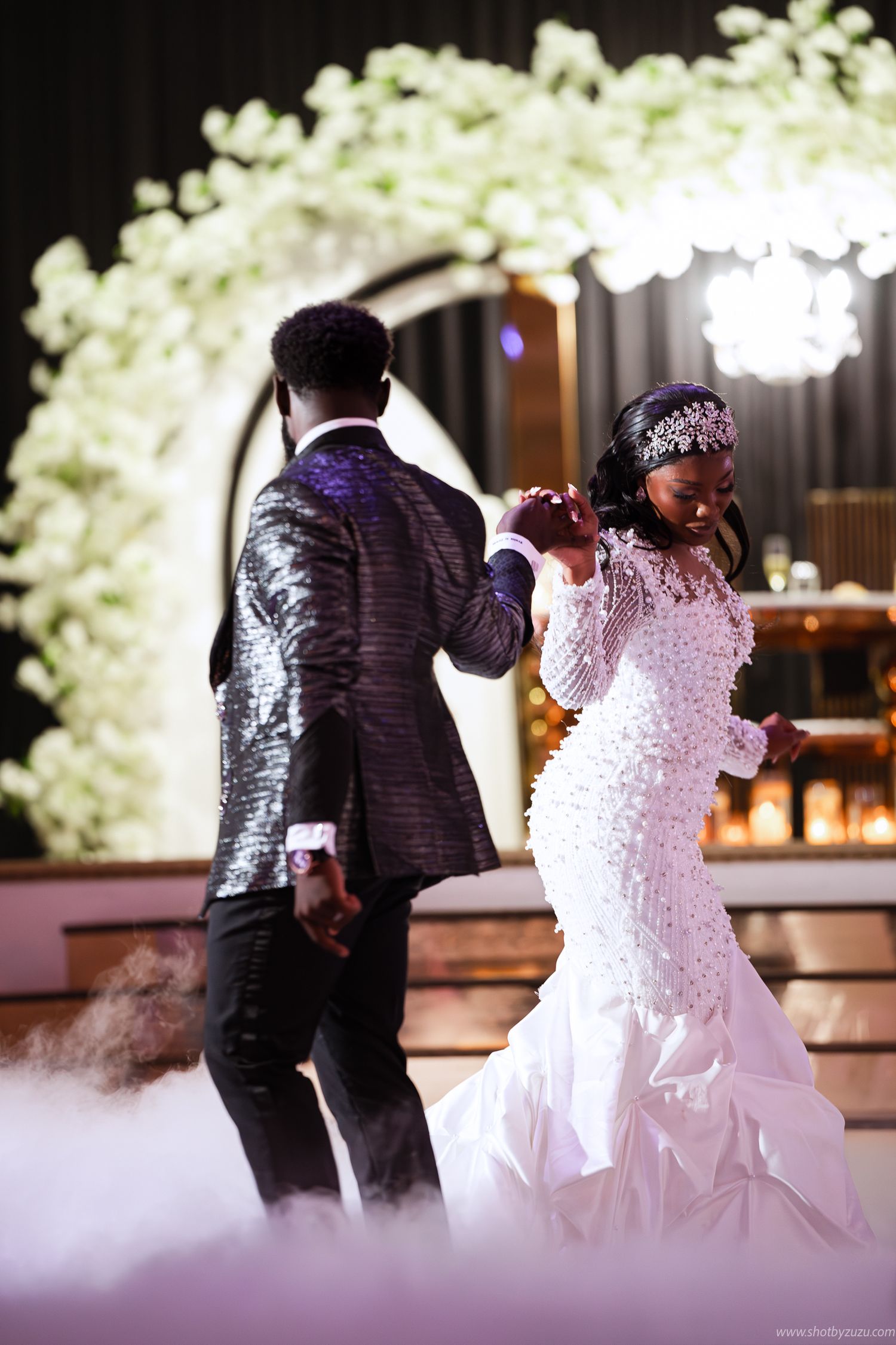 A newlywed couple dances under floral archways with atmospheric fog and romantic lighting at their reception.