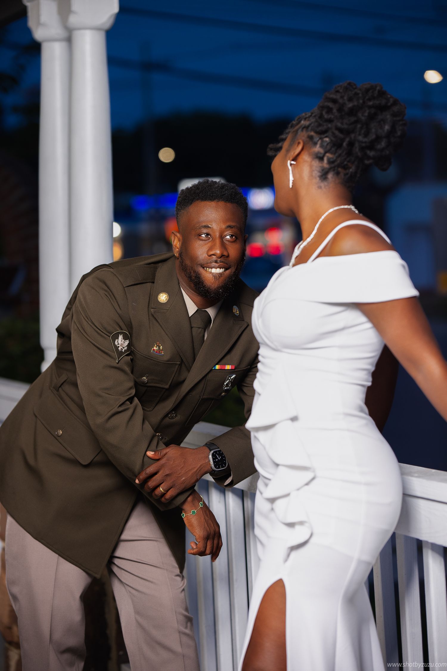 Military groom and bride in elegant white dress share a joyful moment on an outdoor porch at dusk.