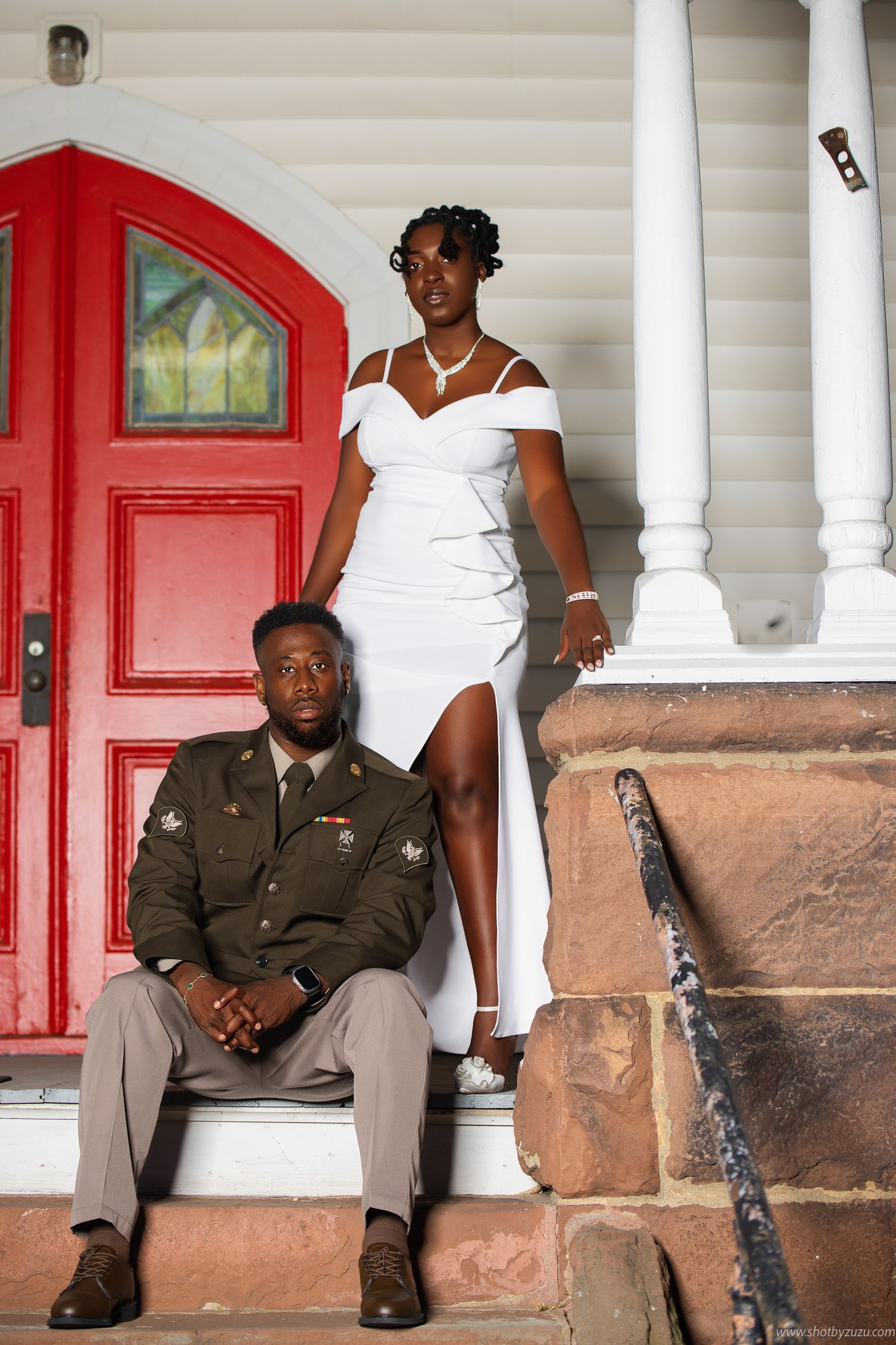 Elegant couple poses on porch steps with classic red door and white columns in formal wedding attire.