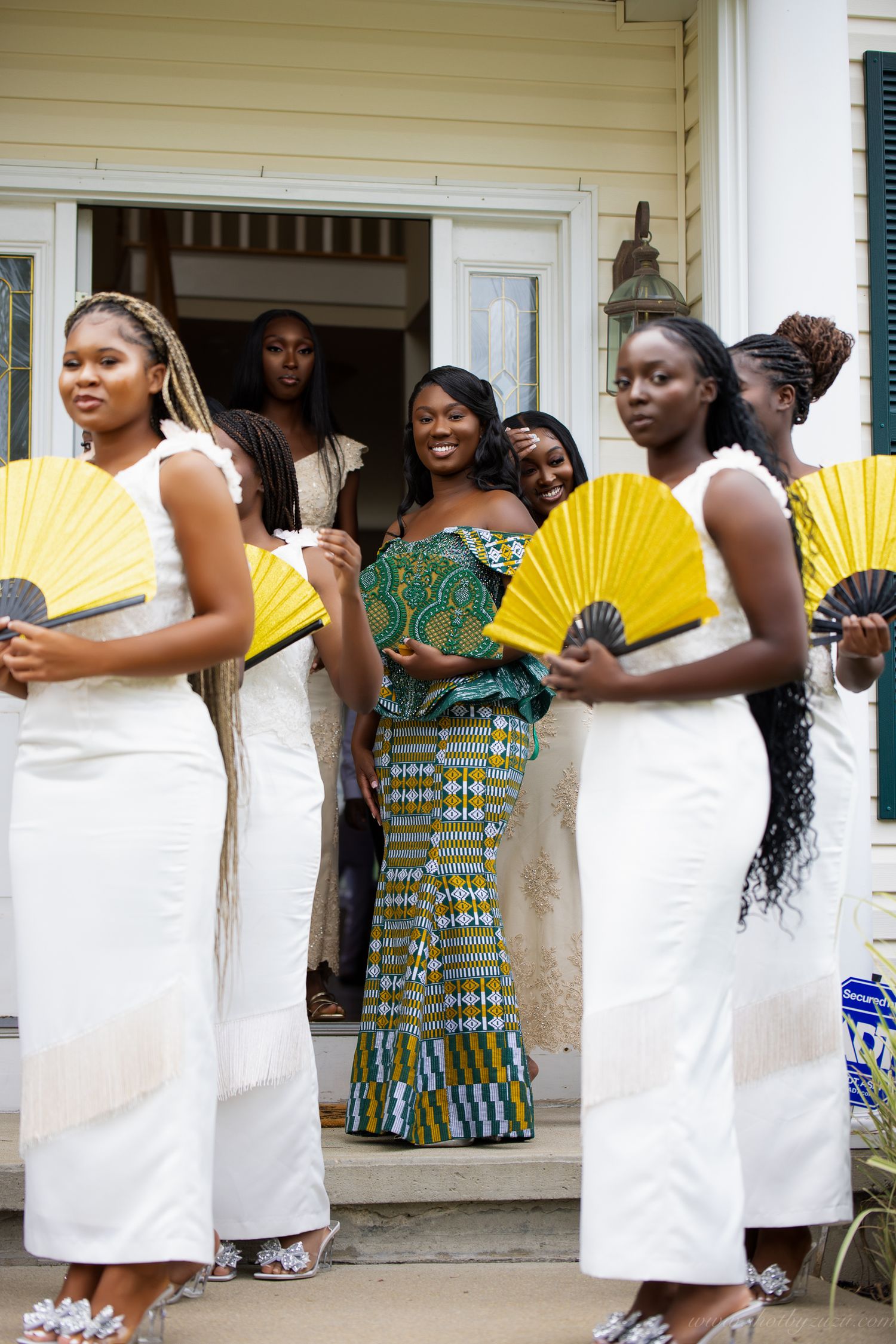 Wedding party poses on porch with yellow fans, wearing white dresses and traditional African attire.