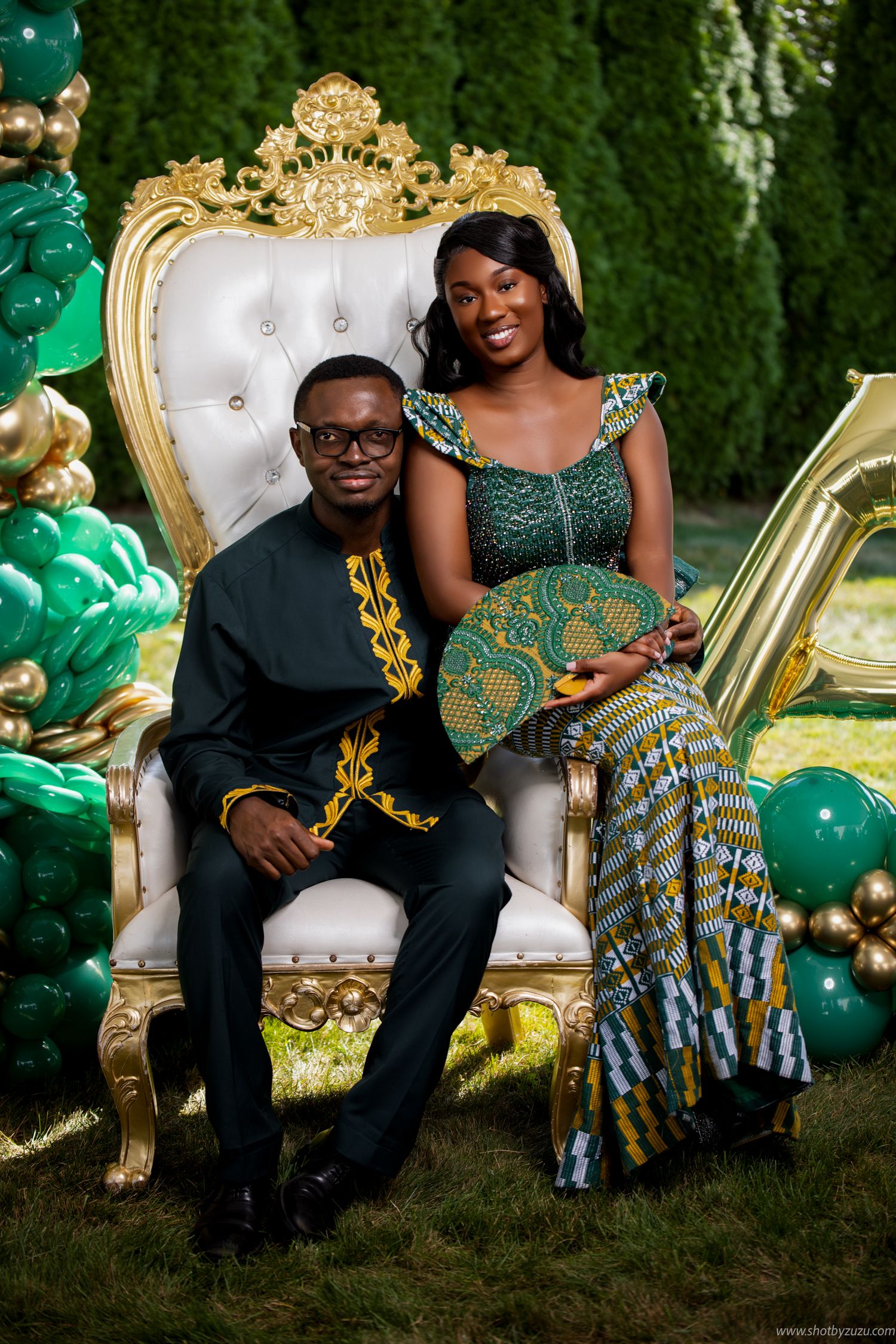 Couple in traditional African attire seated on ornate throne with green and gold decor.