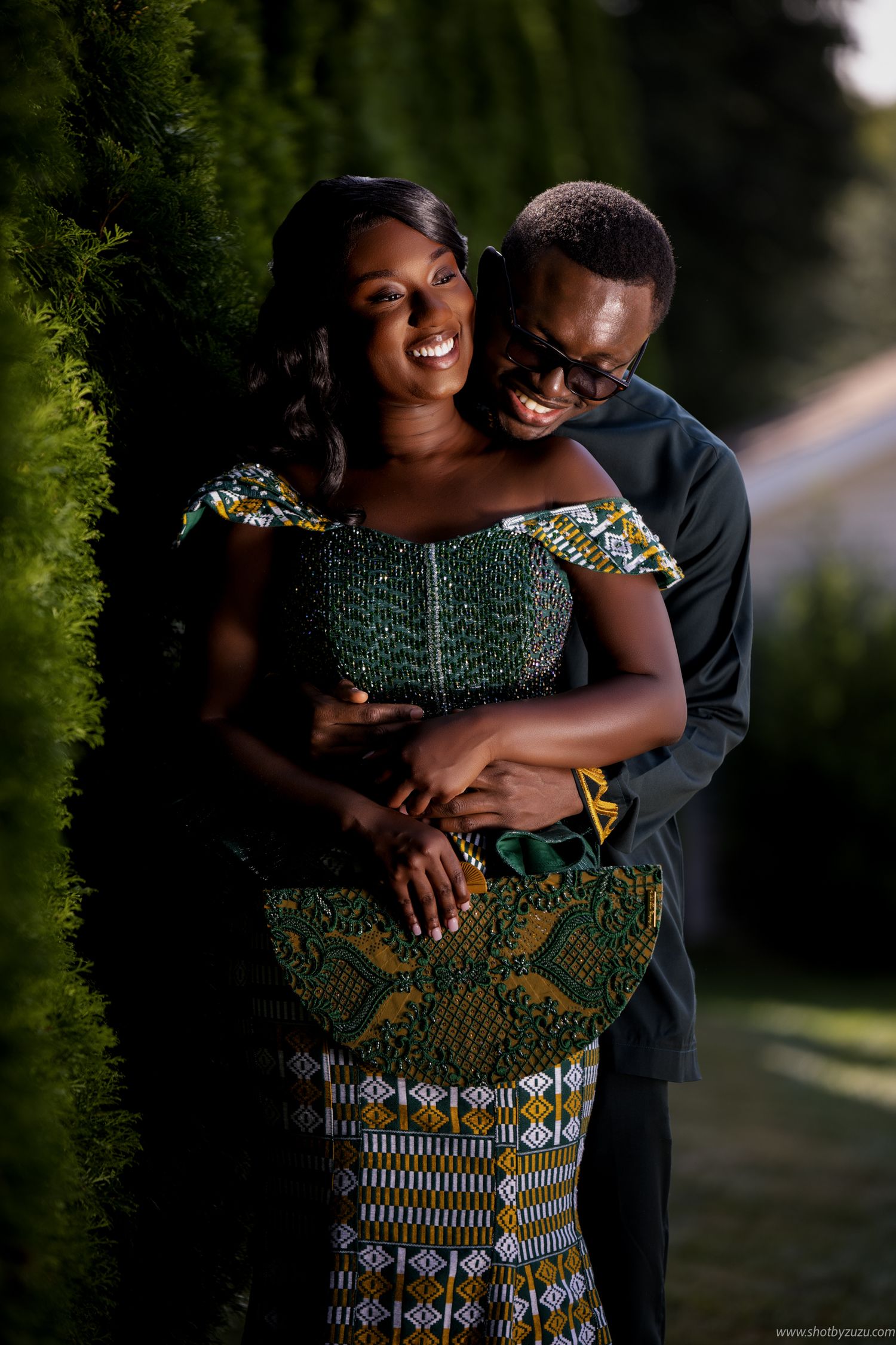 Couple in African dress embracing against green hedge background.