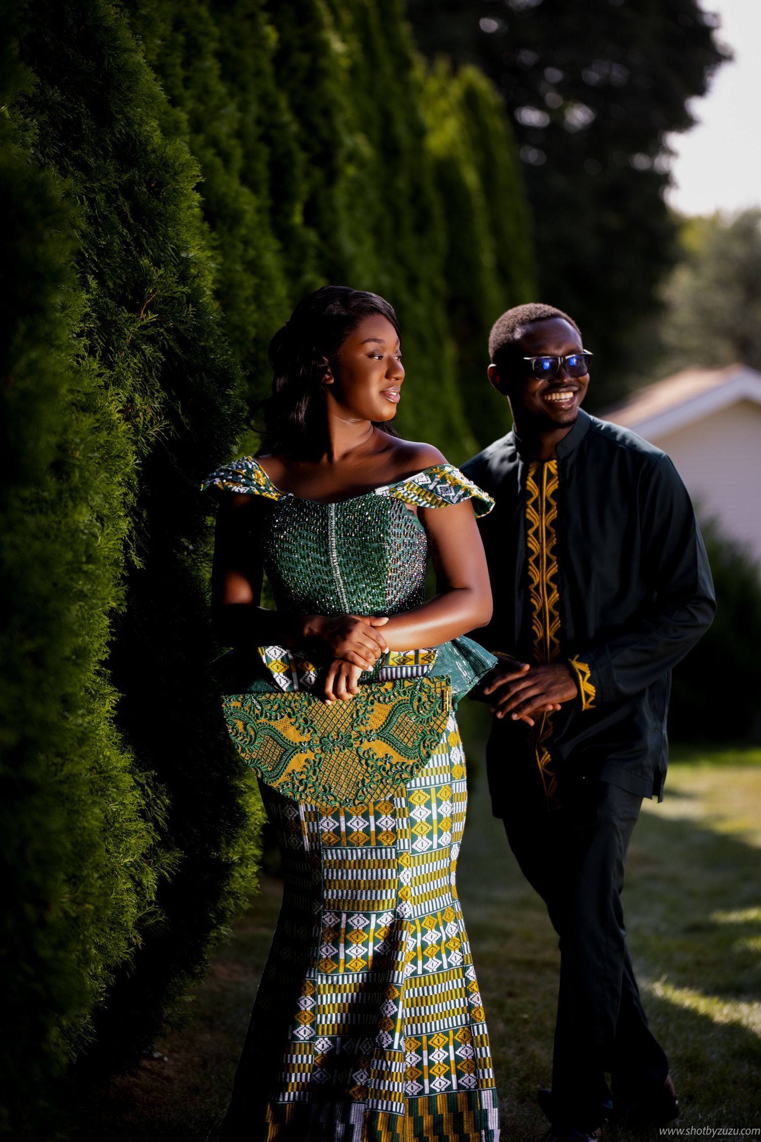 Couple in traditional African attire pose together by hedge at sunset.