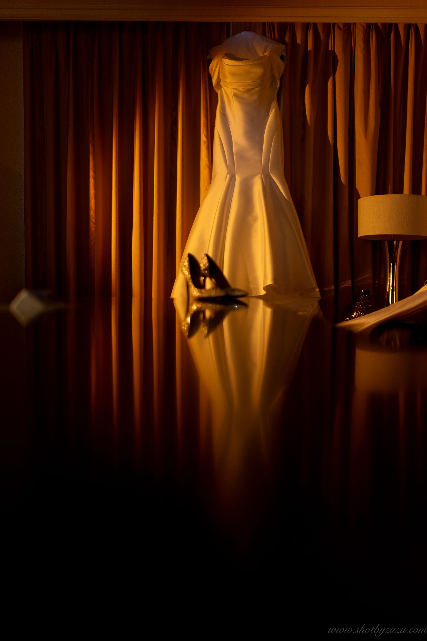 An elegant white wedding dress hangs against warm amber curtains with dramatic lighting.